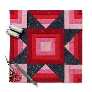 Könnte beinhalten: Ein handgefertigter Quiltblock in Rot-, Rosa- und Dunkelgrautönen mit einem geometrischen Sterndesign. Zwei Spulen grünes Garn, eine Schere und ein Nahttrenner liegen auf weißem Hintergrund.