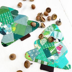 Puede incluir: Dos adornos de árbol de Navidad de tela patchwork verde con bases de fieltro marrón, rodeados de bellotas marrones sobre una superficie blanca.