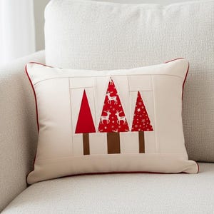 Könnte beinhalten: Rechteckiges cremefarbenes Kissen mit rotem Rand. Es zeigt drei Weihnachtsbaumdesigns in Rot und Braun, wobei ein Baum ein weißes Rentier-Muster aufweist. Das Kissen liegt auf einem weißen Sofa.