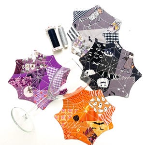 Puede incluir: Posavasos de Halloween con forma de telaraña en telas de patchwork moradas, naranjas y grises. Cada posavasos presenta patrones y diseños únicos, incluyendo fantasmas, murciélagos y figuras de momias. También hay una copa de vino y carretes de hilo.