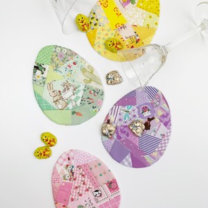 May include: Four colorful, egg-shaped fabric coasters with patchwork designs and Easter-themed illustrations. The coasters are surrounded by clear stemmed glassware and small chocolate Easter bunnies. The coasters are in shades of yellow, green, purple, and pink.
