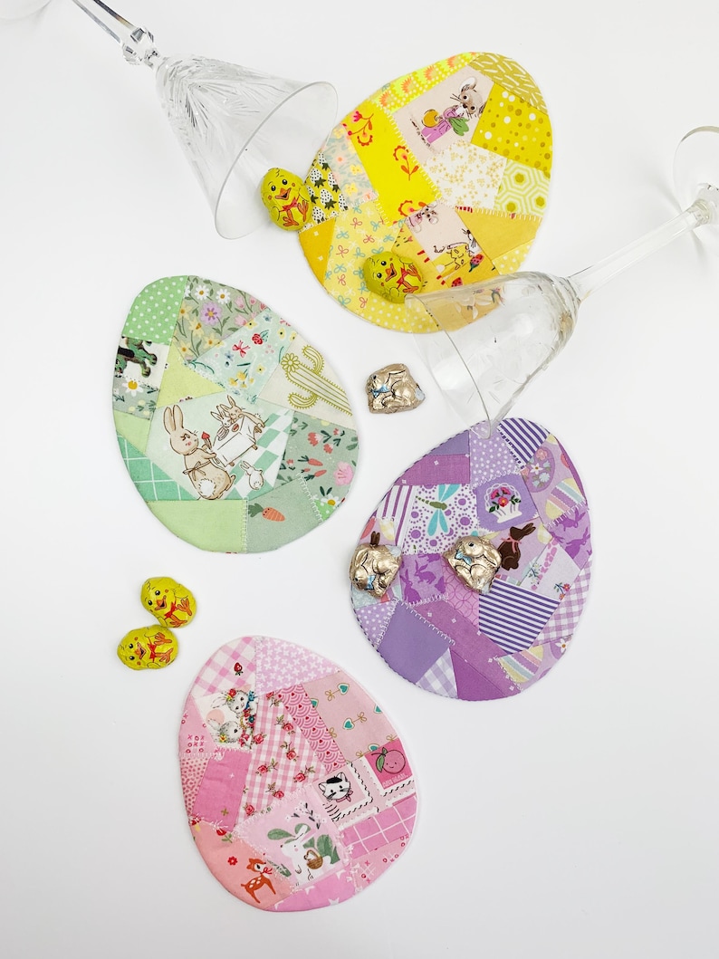 May include: Four colorful, egg-shaped fabric coasters with patchwork designs and Easter-themed illustrations. The coasters are surrounded by clear stemmed glassware and small chocolate Easter bunnies. The coasters are in shades of yellow, green, purple, and pink.