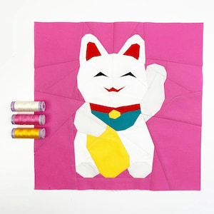 May include: A white origami lucky cat with red ears, a blue collar, and a yellow coin. The cat is sitting on a pink background.