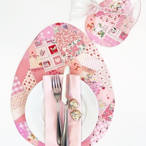 Puede incluir: Un mantel individual ovalado de patchwork rosa y blanco con un plato blanco, una servilleta rosa, un tenedor y un cuchillo de plata, y dos anillos de servilleta de conejo dorado. El mantel individual presenta una variedad de patrones florales y geométricos.
