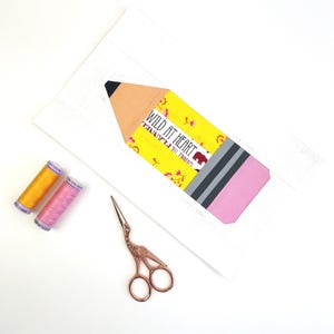 May include: A white rectangular paper featuring a pencil-shaped cutout with a tan tip, a yellow section with the text "WILD AT HEART" and a bear graphic, a gray band, and a pink eraser. Two spools of thread and copper-colored stork scissors.
