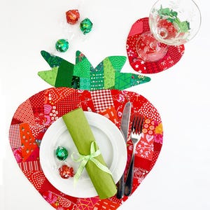 Puede incluir: Un mantel individual de fresa en forma de fresa de color rojo y verde con un plato blanco, una servilleta verde, un tenedor y un cuchillo. También hay tres pequeños chocolates envueltos en verde y rojo sobre la mesa.