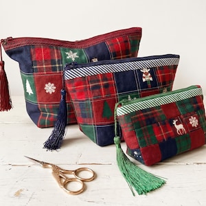 May include: Three zippered pouches in varying sizes made of patchwork fabric featuring red, green, and navy blue plaid squares with embroidered snowflakes, pine trees, and deer motifs. Each pouch includes a long, matching fabric tassel.