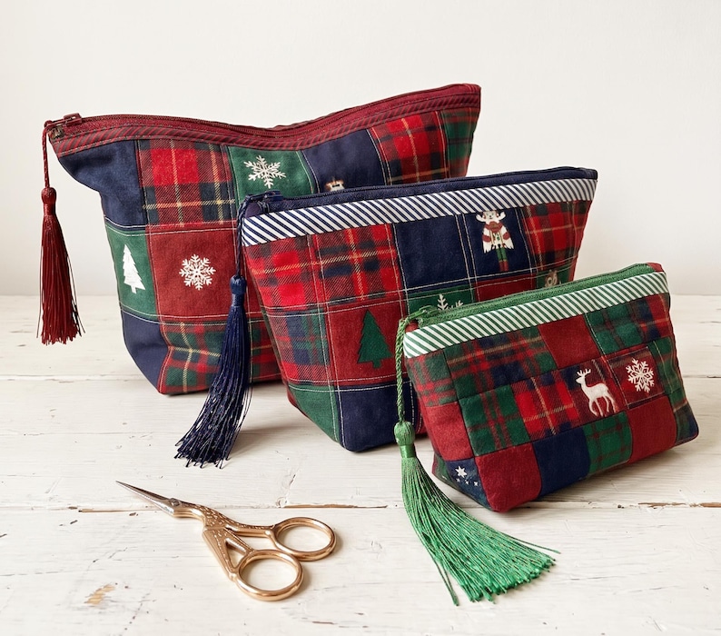 May include: Three zippered pouches in varying sizes made of patchwork fabric featuring red, green, and navy blue plaid squares with embroidered snowflakes, pine trees, and deer motifs. Each pouch includes a long, matching fabric tassel.