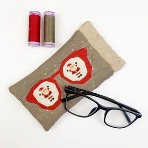May include: A rectangular fabric glasses case featuring a beige background with small gold stars and two red-framed octagonal patches, each displaying a Santa Claus figure. A pair of black-framed eyeglasses and two spools of thread are included.