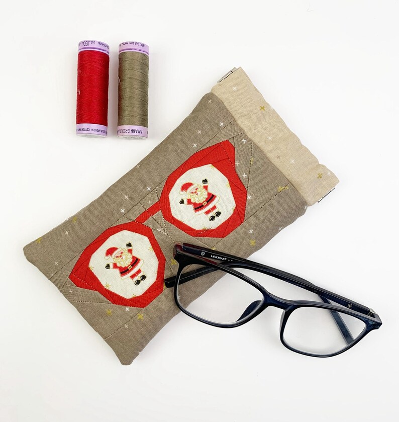 May include: A rectangular fabric glasses case featuring a beige background with small gold stars and two red-framed octagonal patches, each displaying a Santa Claus figure. A pair of black-framed eyeglasses and two spools of thread are included.