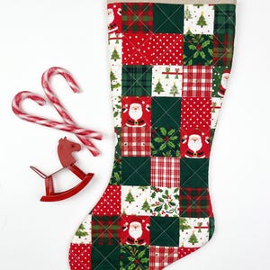 May include: A quilted Christmas stocking featuring a patchwork pattern of red and green squares with prints of Santa Claus, Christmas trees, holly, and plaid designs. Two red and white candy canes and a small red rocking horse ornament.