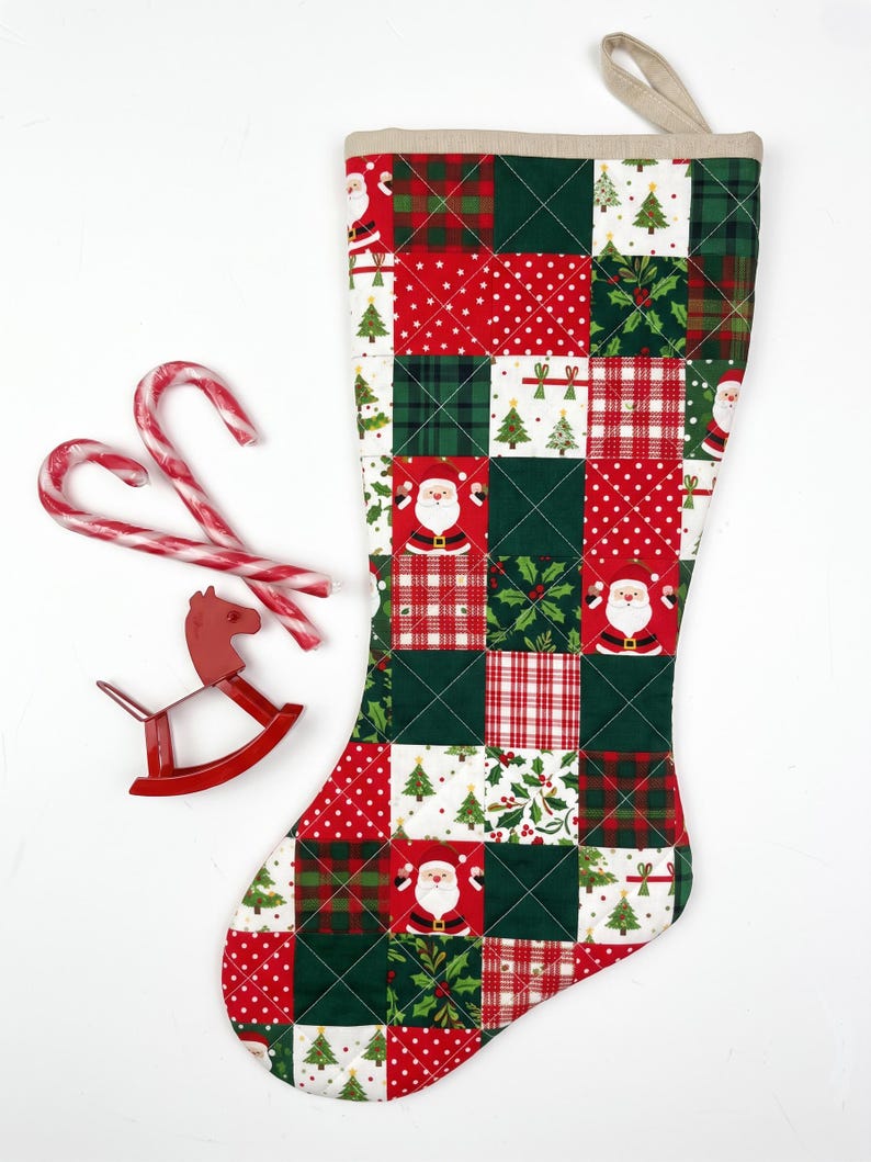 May include: A quilted Christmas stocking featuring a patchwork pattern of red and green squares with prints of Santa Claus, Christmas trees, holly, and plaid designs. Two red and white candy canes and a small red rocking horse ornament.