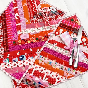 May include: Two red and pink Christmas placemats with a white trim. The placemats are made of fabric scraps sewn together in a patchwork design. One placemat has a candy cane on top of it. The other placemat has a fork and knife on top of it.