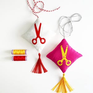 May include: Two fabric sachets with red and yellow tassels. The sachets are shaped like diamonds and have a pair of scissors embroidered on them. One sachet is white with red scissors and the other is pink with yellow scissors. Two spools of thread are also visible in the image.