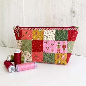May include: A quilted zipper pouch featuring a patchwork design of holiday-themed fabric squares in red, green, pink, and gold, decorated with nutcrackers, gingerbread men, and holly. A silver scissor charm is attached to the zipper pull.