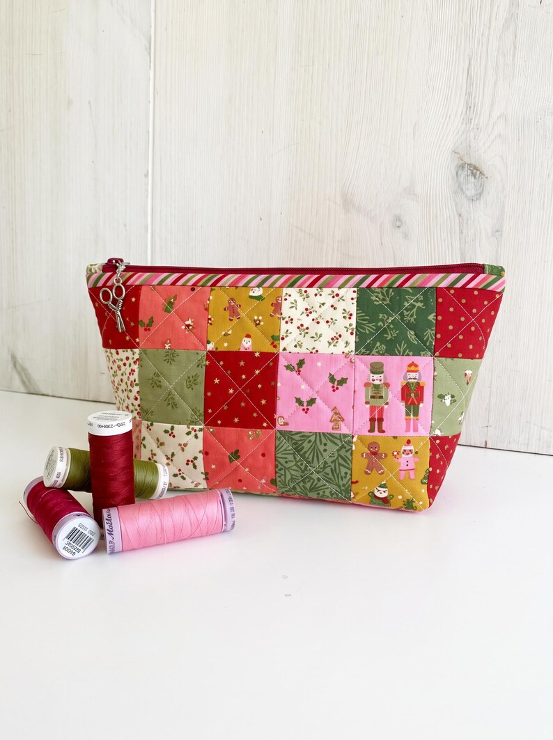 May include: A quilted zipper pouch featuring a patchwork design of holiday-themed fabric squares in red, green, pink, and gold, decorated with nutcrackers, gingerbread men, and holly. A silver scissor charm is attached to the zipper pull.