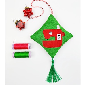 Könnte beinhalten: Ein grüner Stoffschmuck in Form eines Kissens mit einem roten Nähmaschinen-Design. Der Schmuck hat einen grünen Quasten und hängt an einer rot-weißen Schnur. Zwei rote sternförmige Ornamente und zwei Garnrollen, eine rote und eine grüne, sind ebenfalls auf dem Bild zu sehen.