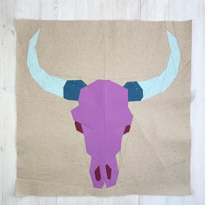 Può includere: Un design patchwork geometrico con un teschio di toro viola con corna azzurre e accenti blu scuro. Il teschio è centrato su un tessuto beige con un sottile motivo a pois. Il design è probabilmente per una trapunta o un arazzo da parete.