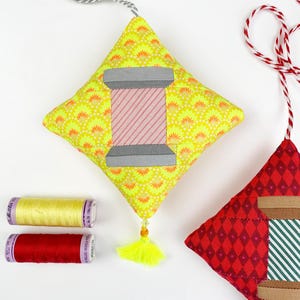 May include: Two handmade fabric ornaments shaped like spools of thread. One ornament is yellow with a pink and grey spool of thread. The other ornament is red with a green and brown spool of thread. Both ornaments have a tassel.