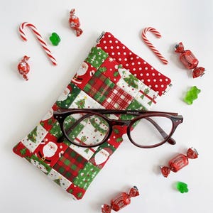 May include: A quilted fabric glasses case featuring a patchwork pattern of Santa Claus, Christmas trees, and red plaid squares, topped with a red and white polka dot border. A pair of dark brown tortoiseshell eyeglasses rests on the case.
