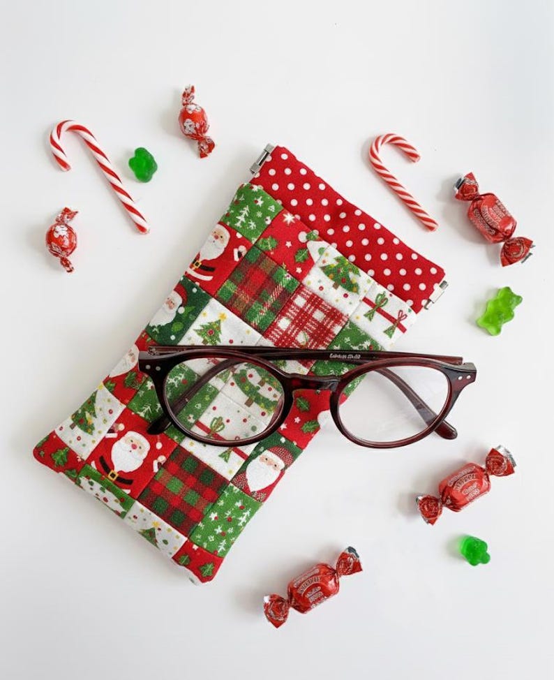 May include: A quilted fabric glasses case featuring a patchwork pattern of Santa Claus, Christmas trees, and red plaid squares, topped with a red and white polka dot border. A pair of dark brown tortoiseshell eyeglasses rests on the case.
