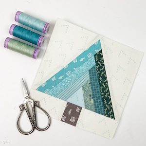 May include: A close-up of a quilt block with a geometric design in shades of teal, green, and brown. The block is made of various fabrics with different patterns, including stripes, polka dots, and floral prints. The block is surrounded by a white background with a pattern of small triangles.