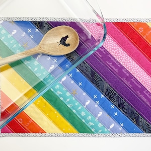 Puede incluir: Un salvamanteles acolchado de colores arcoíris con un borde blanco y una cuchara de madera con una silueta de gallo en la parte superior. El salvamanteles está hecho de tiras de tela con diferentes patrones y colores, incluyendo rojo, naranja, amarillo, verde, azul, morado y rosa. El texto "hermosa luchadora creativa empresaria madura diosa educada" es visible en la tira de tela morada.