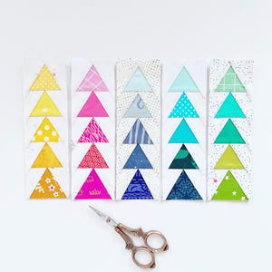 May include: A set of six fabric triangles arranged in a row, each triangle is made of a different patterned fabric. The fabrics are yellow, pink, blue, turquoise, green, and lime green. A pair of silver scissors is in the foreground.