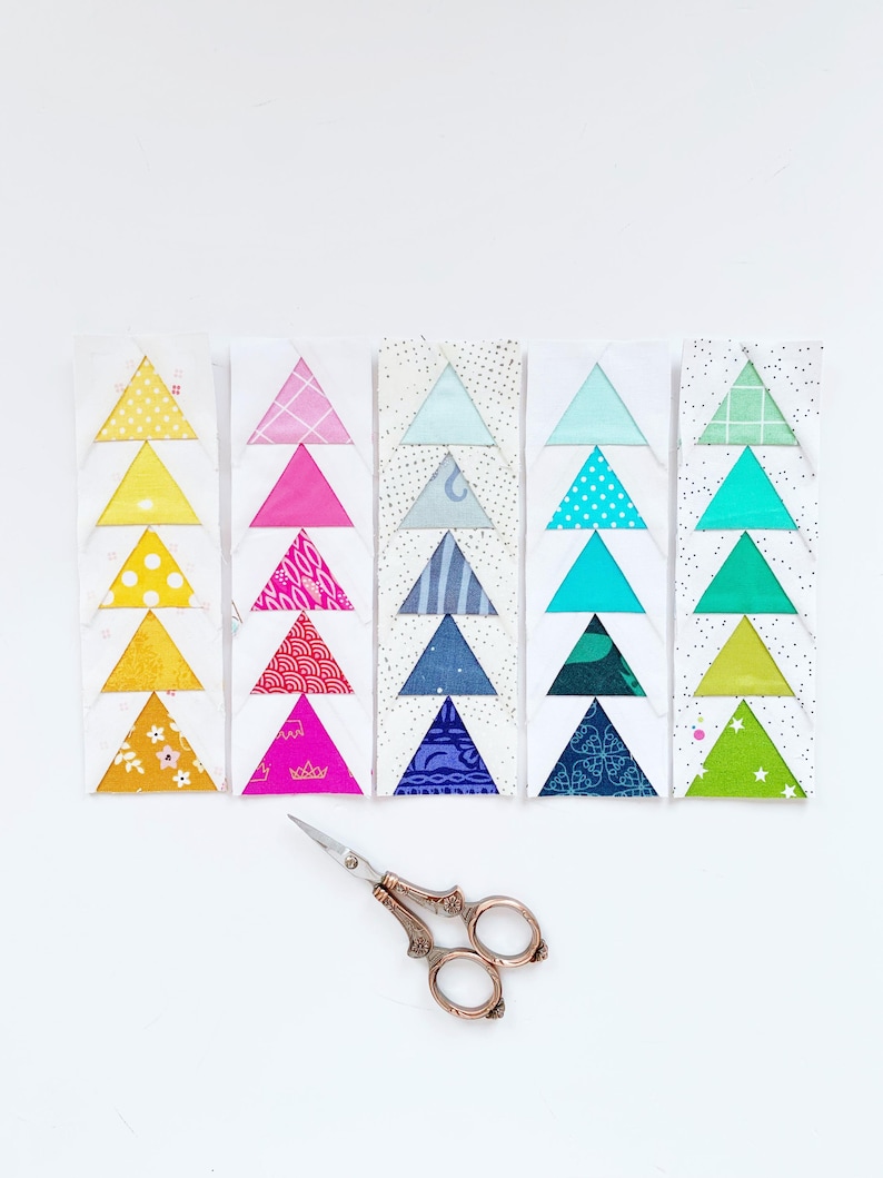 May include: A set of six fabric triangles arranged in a row, each triangle is made of a different patterned fabric. The fabrics are yellow, pink, blue, turquoise, green, and lime green. A pair of silver scissors is in the foreground.