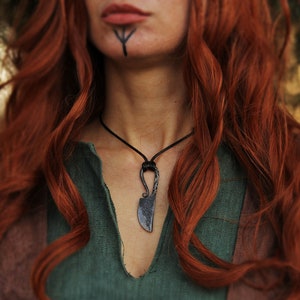 Puede incluir: Una mujer con el pelo largo y rojo lleva una camisa de lino verde y un collar con un pequeño colgante de cuchillo de metal negro.