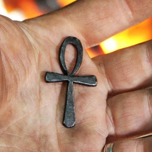 May include: A black metal Ankh pendant, a symbol of life and immortality in ancient Egypt, is held in a hand against a blurry background of fire.