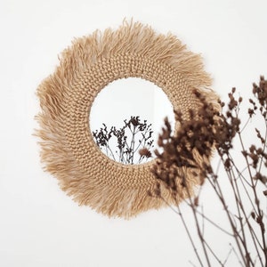 May include: A round mirror with a woven jute frame and a fringe border. The mirror is hanging on a white wall and reflects dried flowers.