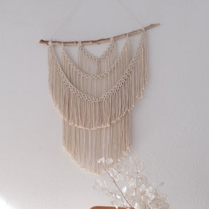 Könnte beinhalten: Ein beigefarbener Makramee-Wandbehang mit einem natürlichen Ast aus Holz oben. Der Behang besteht aus geknüpften Schnüren und hat unten einen Fransen. Der Behang wird an einer weißen Wand mit einer Vase mit Trockenblumen davor gezeigt.