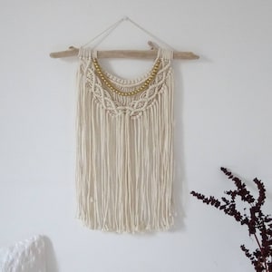 May include: A white macrame wall hanging with a gold beaded trim, hanging from a driftwood branch.