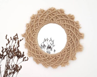 May include: A round mirror with a macrame frame made of natural jute rope. The frame has a fringe detail and a mandala pattern. The mirror is reflecting a branch with dried flowers.