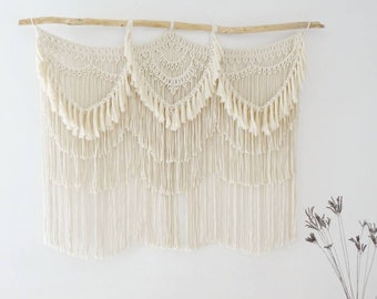 May include: A white macrame wall hanging with three sections, each featuring a crescent moon shape with fringe details. The hanging is attached to a natural wood branch.