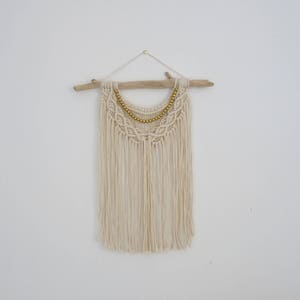 May include: A white macrame wall hanging with a wooden dowel and gold beads. The hanging has a fringe design and is perfect for adding a bohemian touch to any room.