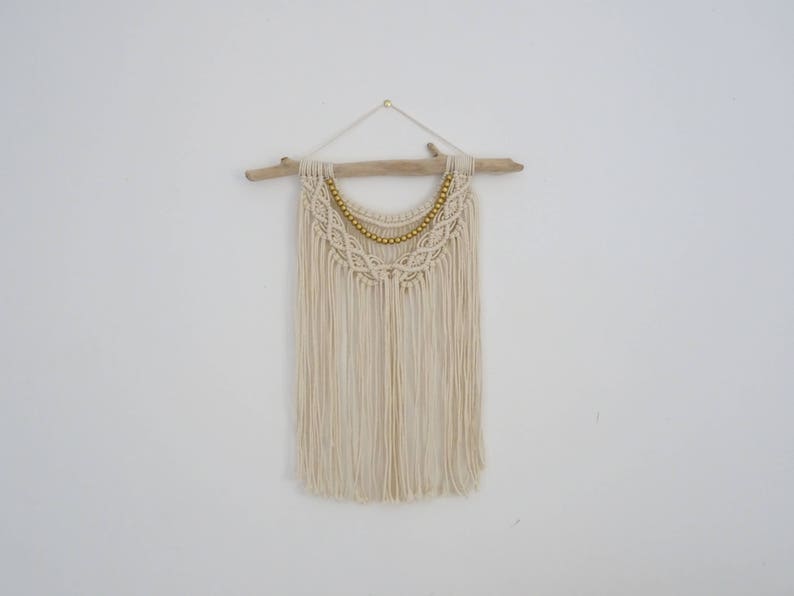 May include: A white macrame wall hanging with a wooden dowel and gold beads. The hanging has a fringe design and is perfect for adding a bohemian touch to any room.