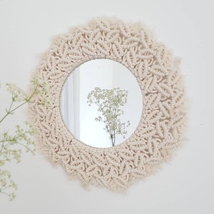 May include: A round mirror with a macrame frame in a cream color. The mirror is hanging on a white wall.