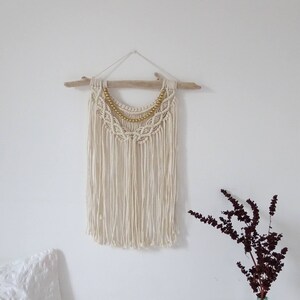 May include: A white macrame wall hanging with a gold beaded trim. The hanging is attached to a piece of driftwood.
