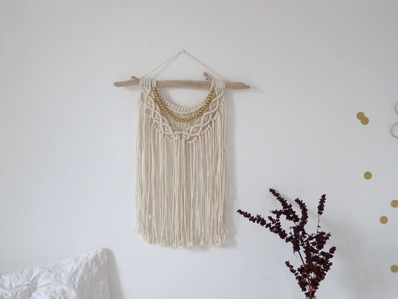 May include: A white macrame wall hanging with a gold beaded trim. The hanging is attached to a piece of driftwood.