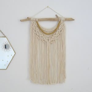 May include: A white macrame wall hanging with a driftwood dowel and gold beads. The hanging has a fringe of white yarn.
