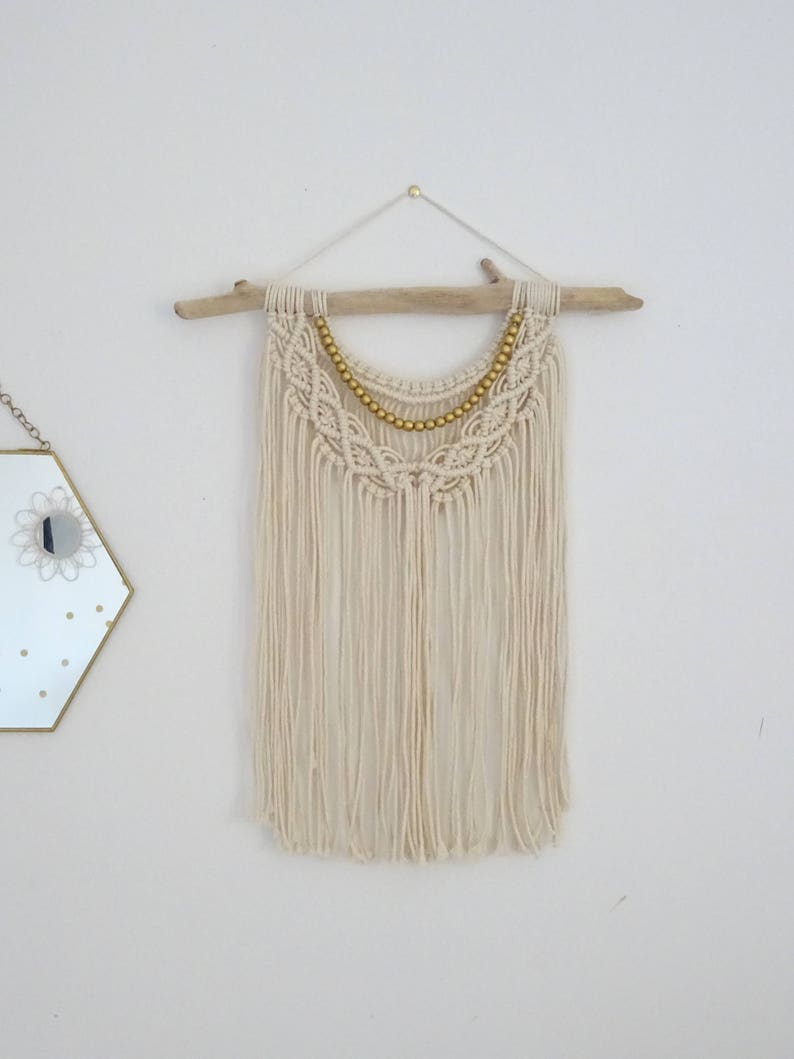 May include: A white macrame wall hanging with a driftwood dowel and gold beads. The hanging has a fringe of white yarn.