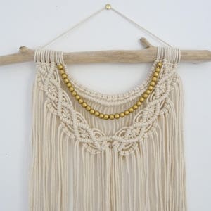 May include: A white macrame wall hanging with a gold beaded trim. The hanging is attached to a driftwood branch.