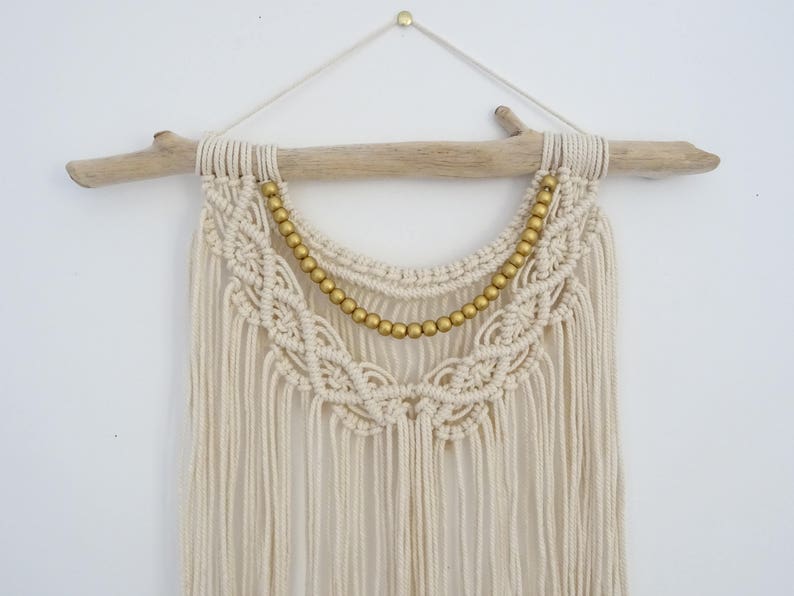 May include: A white macrame wall hanging with a gold beaded trim. The hanging is attached to a driftwood branch.