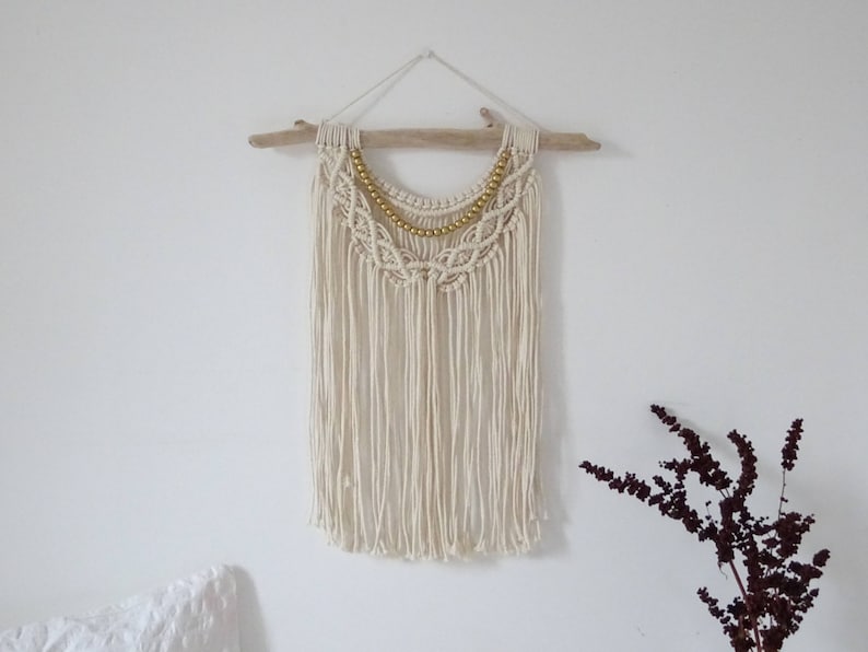 May include: A white macrame wall hanging with a gold beaded trim, hanging from a driftwood branch.