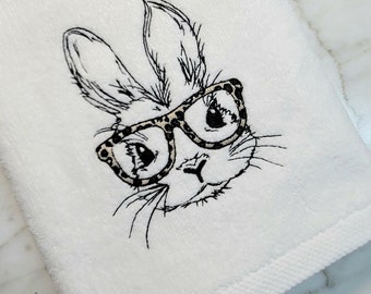 May include: White hand towel with an embroidered bunny wearing glasses with a leopard print pattern. The bunny's face is detailed with black thread, creating a whimsical design. The towel is made of a soft, absorbent material.