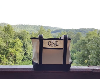 May include: A white canvas tote bag with black trim and handles. The bag has a black embroidered monogram "CNL" on the front. The bag is sitting on a wooden railing with a view of a green forest in the background.