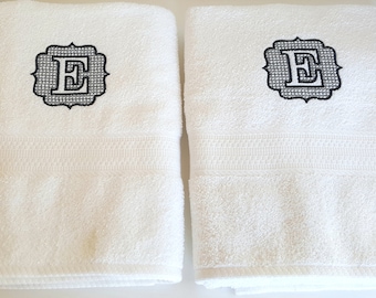 May include: Two white bath towels with black embroidered monograms in the center of each towel. The monogram is a stylized letter "E" with a diamond pattern inside the letter.