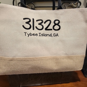 May include: A beige canvas tote bag with black trim and a black embroidered design that says "31328 Tybee Island, GA".